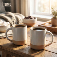 Two Dove Grey stoneware mugs with coffee on a wooden table by a window
