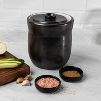 Black glazed 7L fermentation crock on a marble counter with cabbage, cucumbers, garlic and pink salt