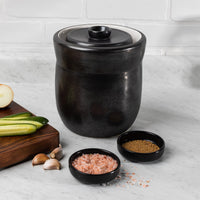 Black glazed 3L fermentation crock on a marble counter with cabbage, cucumbers, garlic and pink salt