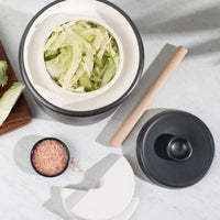 Overhead view of the 7L crock filled with cabbage, with wooden tamper and ceramic weights on marble