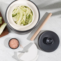 Overhead view of the 3L crock filled with cabbage, with wooden tamper and ceramic weights on marble